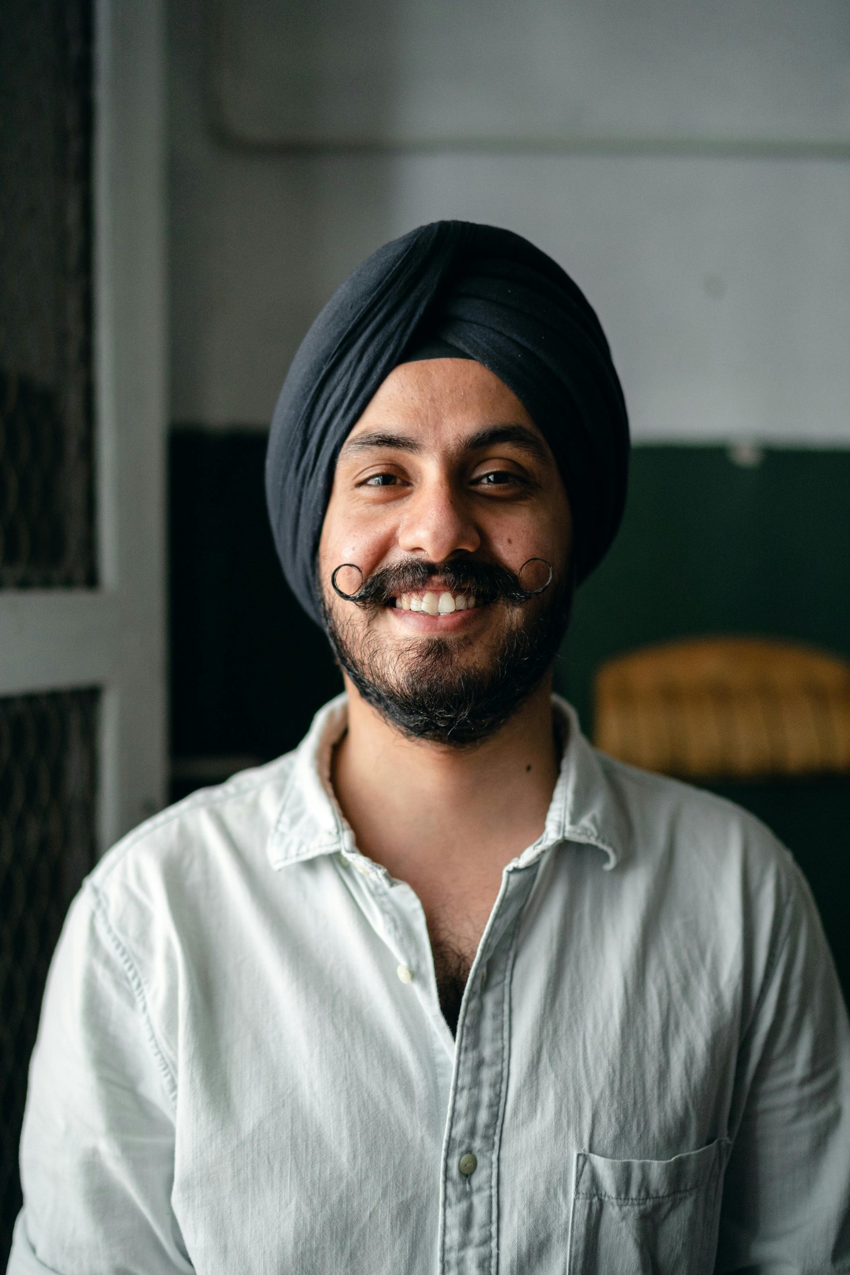Positive bearded Indian male in shirt and turban standing in room and smiling while looking at camera